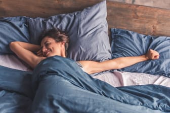 Eine Frau streckt sich entspannt im Bett: Wer leichte Einschlafprobleme hat, kann von der 4-7-8-Atmung wahrscheinlich profitieren.