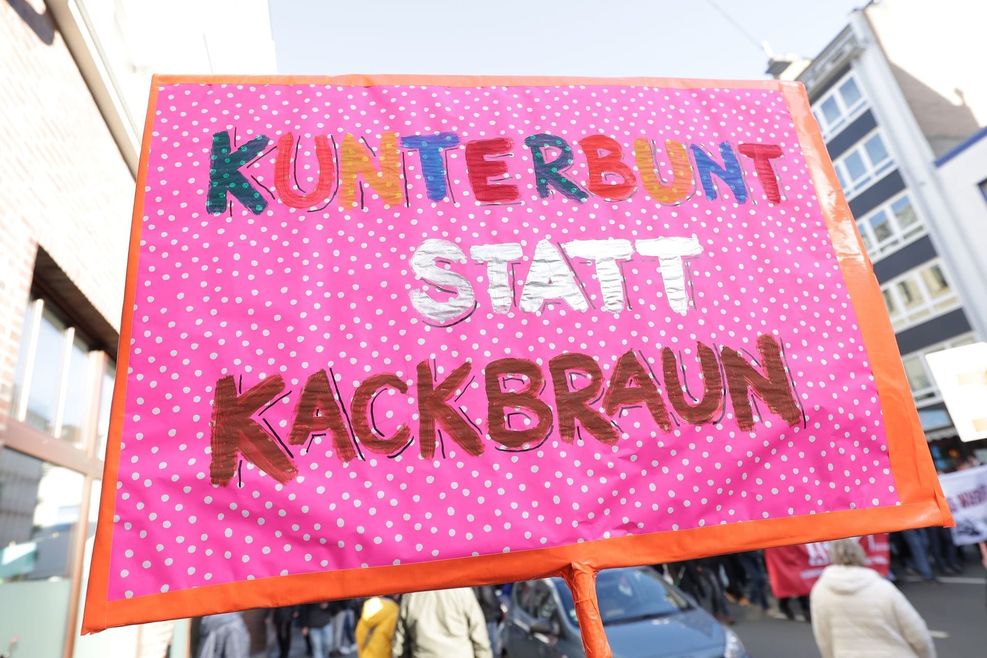 "Kunterbunt statt kackbraun" steht auf einem rosa Plakat mit weißen Punkten, das auf einer Demonstration gegen rechts zu sehen.