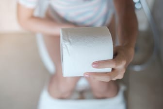 Eine auf der Toilettenschüssel sitzende Frau hält eine Rolle Toilettenpapier in der Hand.