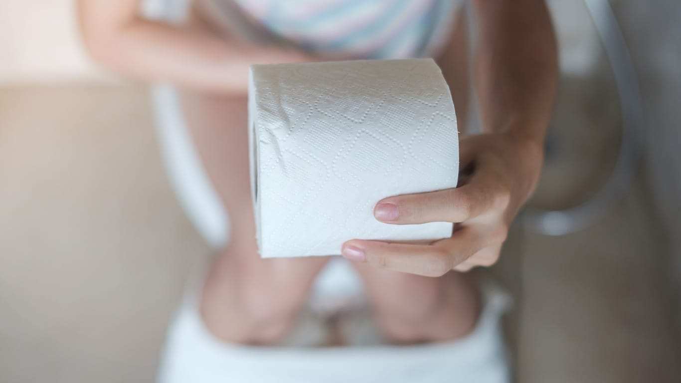 Eine auf der Toilettenschüssel sitzende Frau hält eine Rolle Toilettenpapier in der Hand.