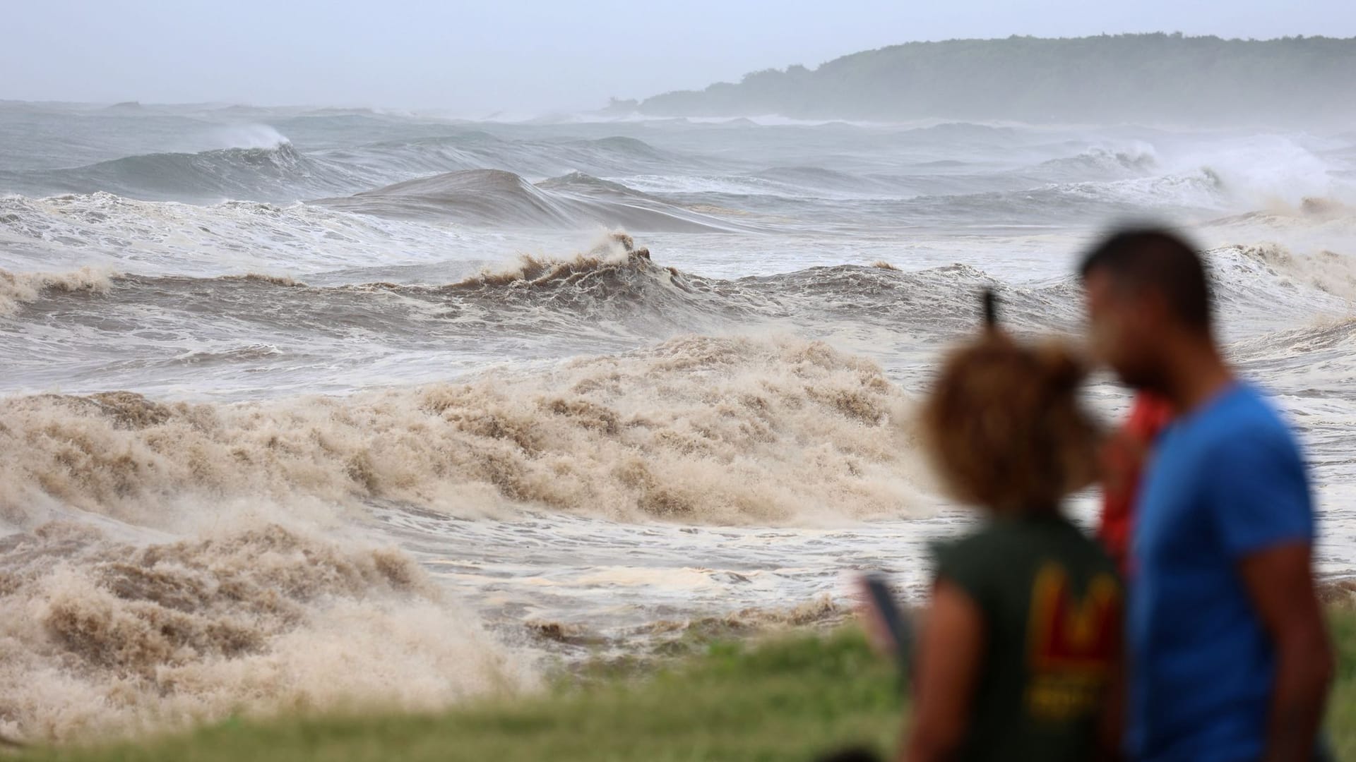 La Réunion: Zwei Personen fotografieren die hohen Wellen von der Strandpromenade aus.