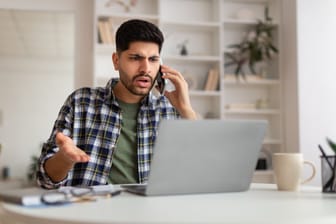 Ein Mann sitzt am Schreibtisch vor einem Laptop. Er hat ein Telefon am Ohr und schaut verärgert.