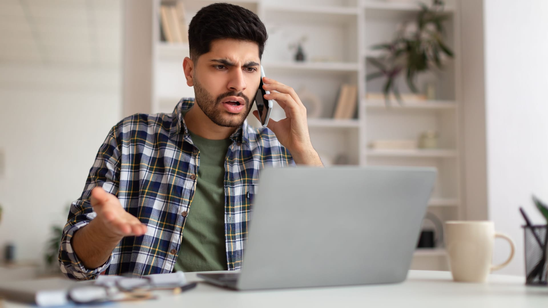 Ein Mann sitzt am Schreibtisch vor einem Laptop. Er hat ein Telefon am Ohr und schaut verärgert.