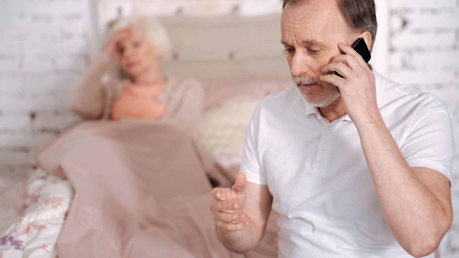 Mann am Telefon, im Hintergrund eine kranke ältere Frau im Bett