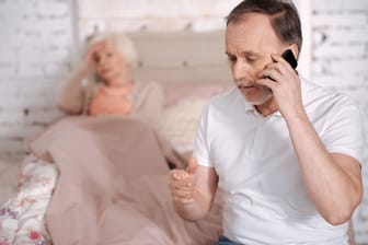 Mann am Telefon, im Hintergrund eine kranke ältere Frau im Bett
