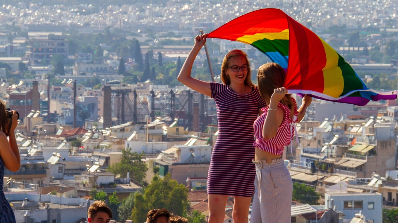 Girls holding flag on Athens Pride 2018 day being photographed by their friends on a rock accross the Acropolis as other tourists look