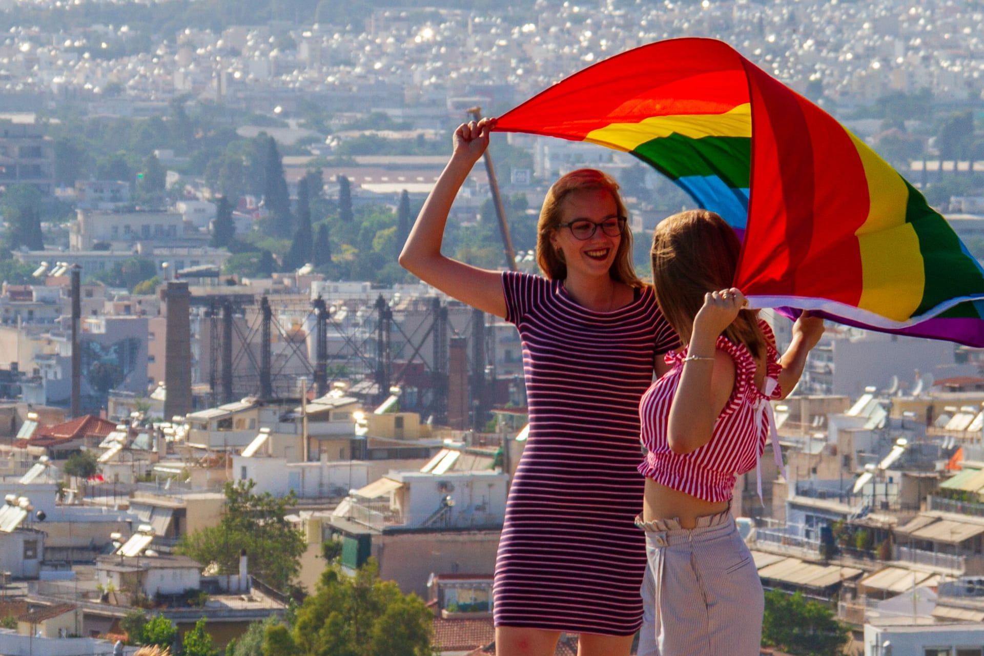 Girls holding flag on Athens Pride 2018 day being photographed by their friends on a rock accross the Acropolis as other tourists look