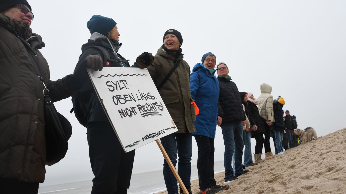 Einheimische und Feriengäste Hand in Hand am Strand auf Sylt: Auf dem Plakat steht "Sylt. Oben links. Nicht rechts!"