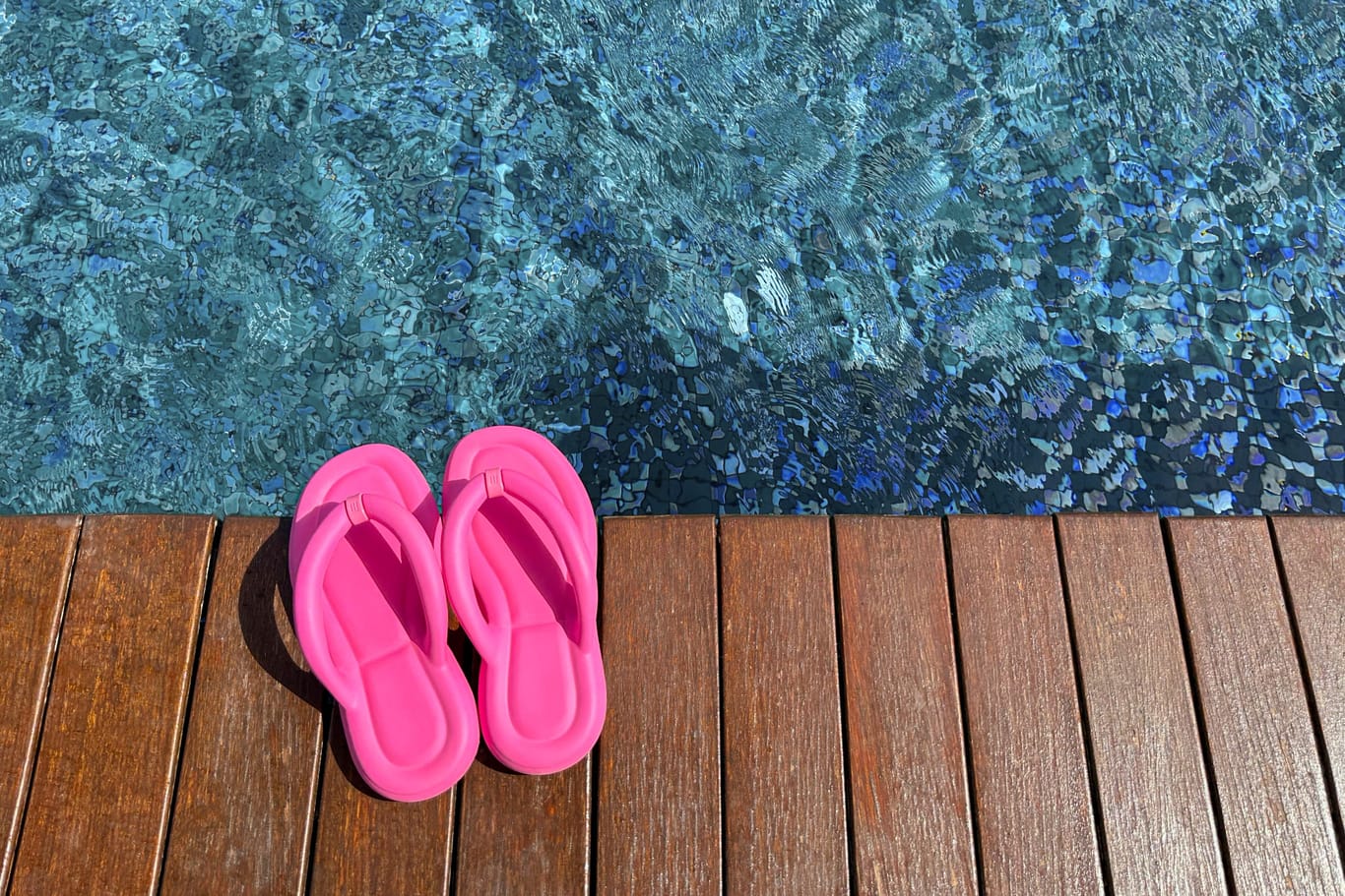 Clear rippled water in swimming pool and pink flip-flops on wooden deck outdoors, above view. Space for text