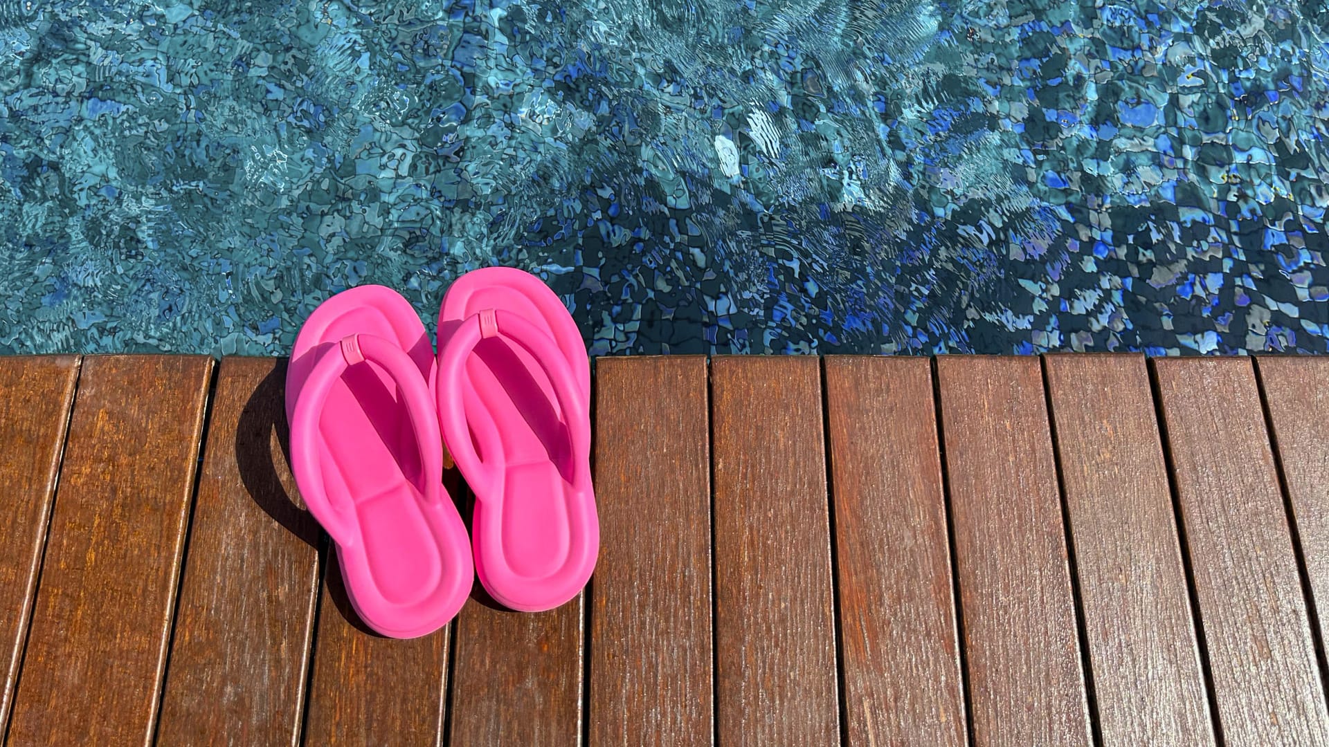 Clear rippled water in swimming pool and pink flip-flops on wooden deck outdoors, above view. Space for text