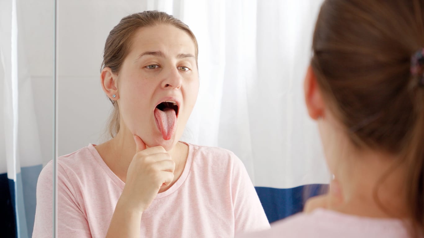 Frau prüft im Spiegel ihre Zunge: Das Sjögren-Syndrom wirkt sich auf vielfältige Weise auf den Körper aus, häufig auch auf die Zunge.
