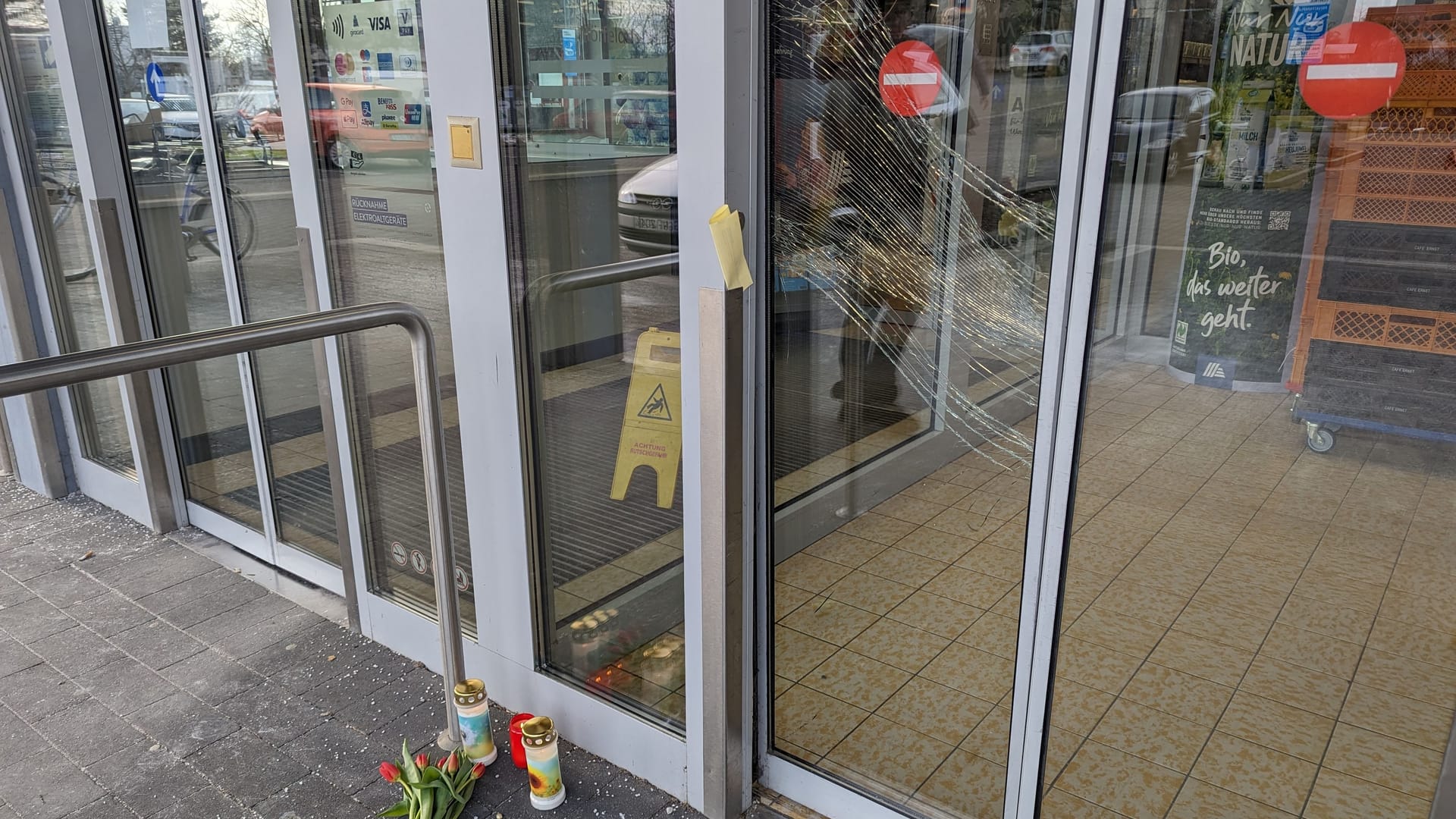 Blumen und Kerzen vor der Aldi-Filale in Mörfelden-Walldorf. Zu sehen ist auch die eingetretene Tür zur Filale, die Augenzeugenberichten zufolge versperrt und gewaltsam wieder aufgebrochen wurde.