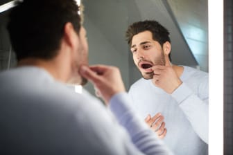 Ein Mann prüft seine Zähne im Spiegel: Zahnstein und Karies können Zähne und Zahnfleisch gefährden.