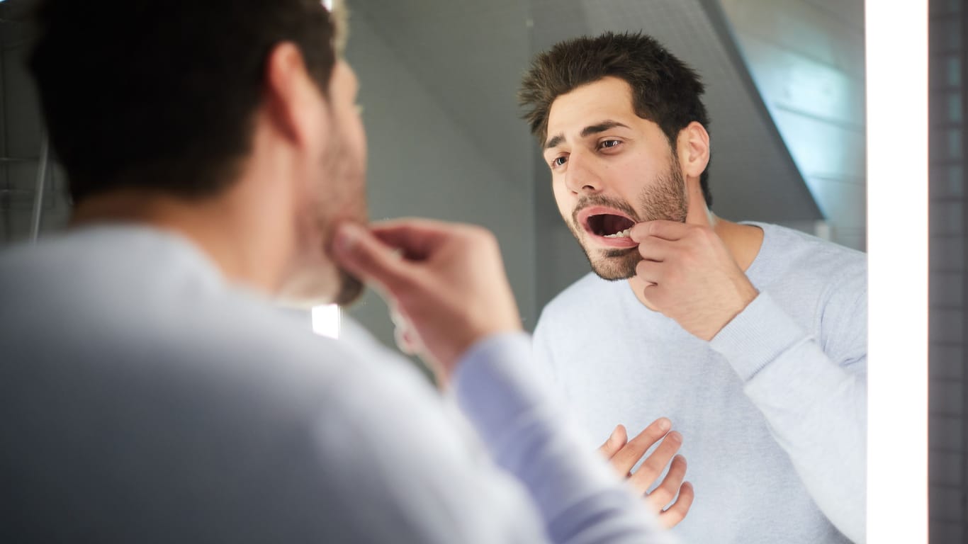 Ein Mann prüft seine Zähne im Spiegel: Zahnstein und Karies können Zähne und Zahnfleisch gefährden.