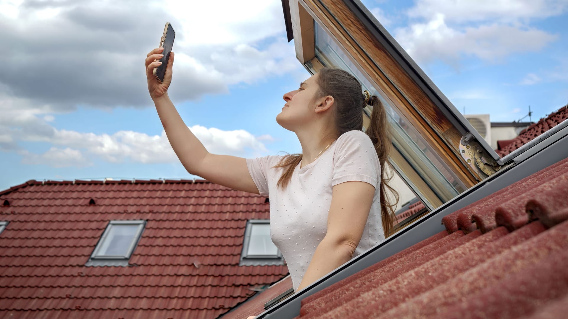 Young woman experiences signal issues with her cellphone as she peers out of an open attic window, attempting to regain a connection. Frustration of lost signal and connectivity problems