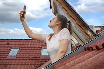 Young woman experiences signal issues with her cellphone as she peers out of an open attic window, attempting to regain a connection. Frustration of lost signal and connectivity problems