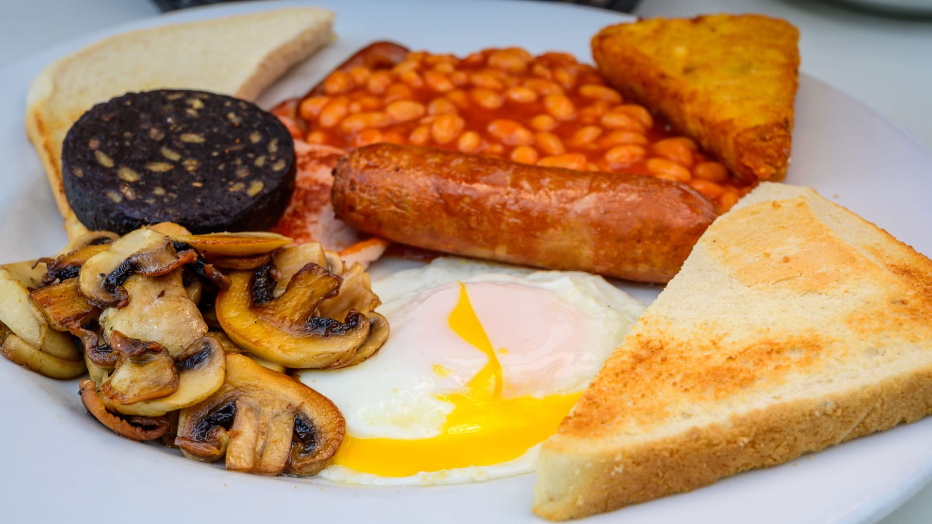 White board with full English breakfast with bacon, fried egg, beans, tomato, roasted sausage, black pudding, scons, hash browns and fried mushrooms