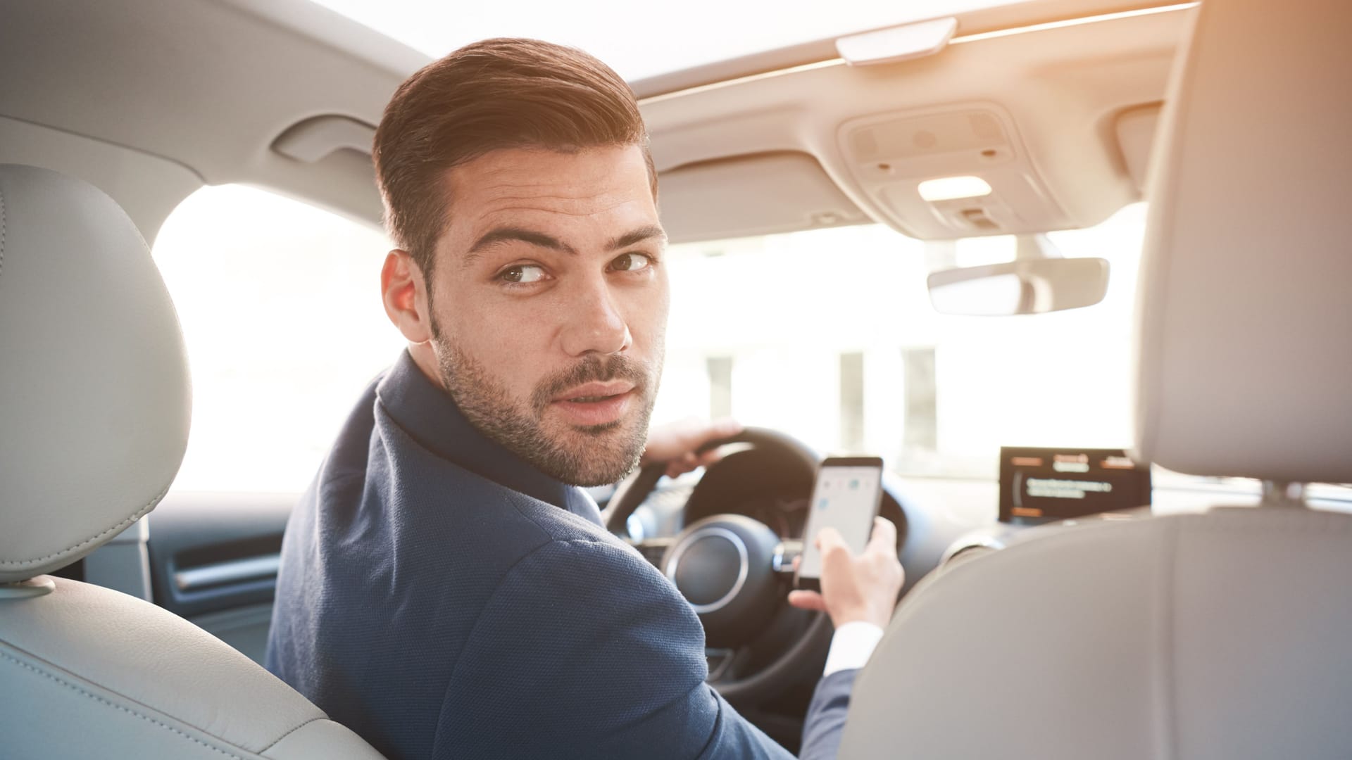 Portrait of stylish confident businessman in car wearing suit, turning to back seats with serious expression.