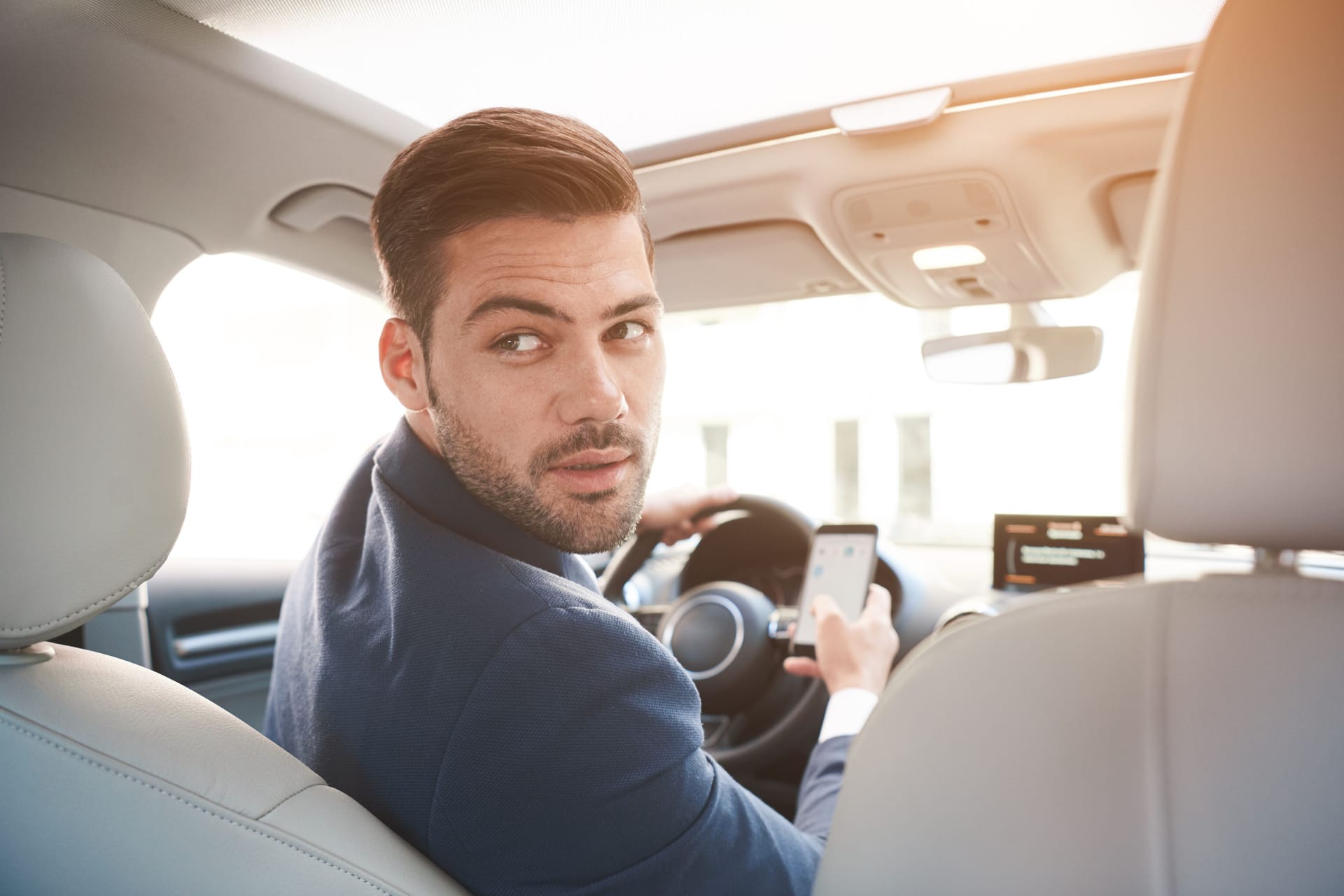 Portrait of stylish confident businessman in car wearing suit, turning to back seats with serious expression.