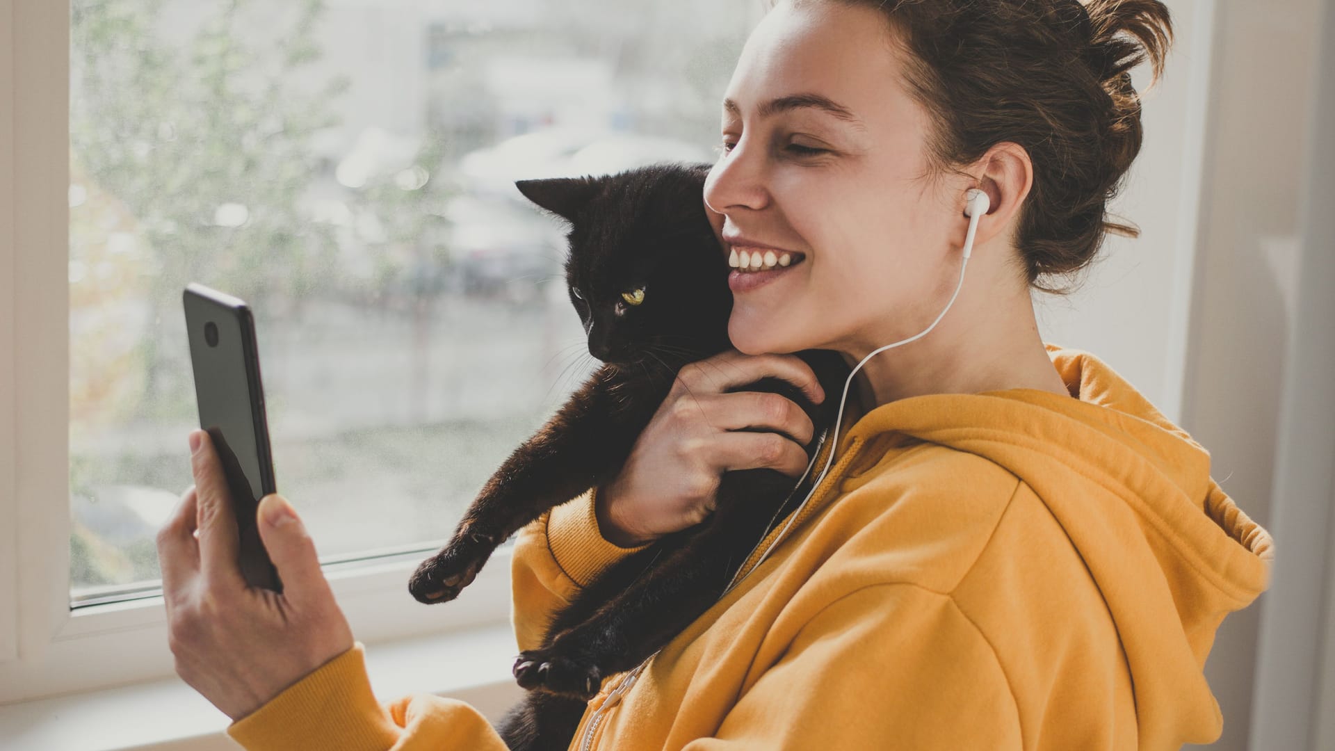 Cheerful young woman wearing headphones holds black pet cat using smartphone for video call, gesturing hi to friend or parent.Caucasian girl in yellow hoodie making selfie,sharing data on social