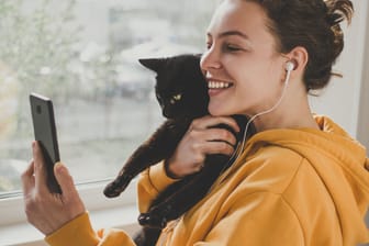 Cheerful young woman wearing headphones holds black pet cat using smartphone for video call, gesturing hi to friend or parent.Caucasian girl in yellow hoodie making selfie,sharing data on social