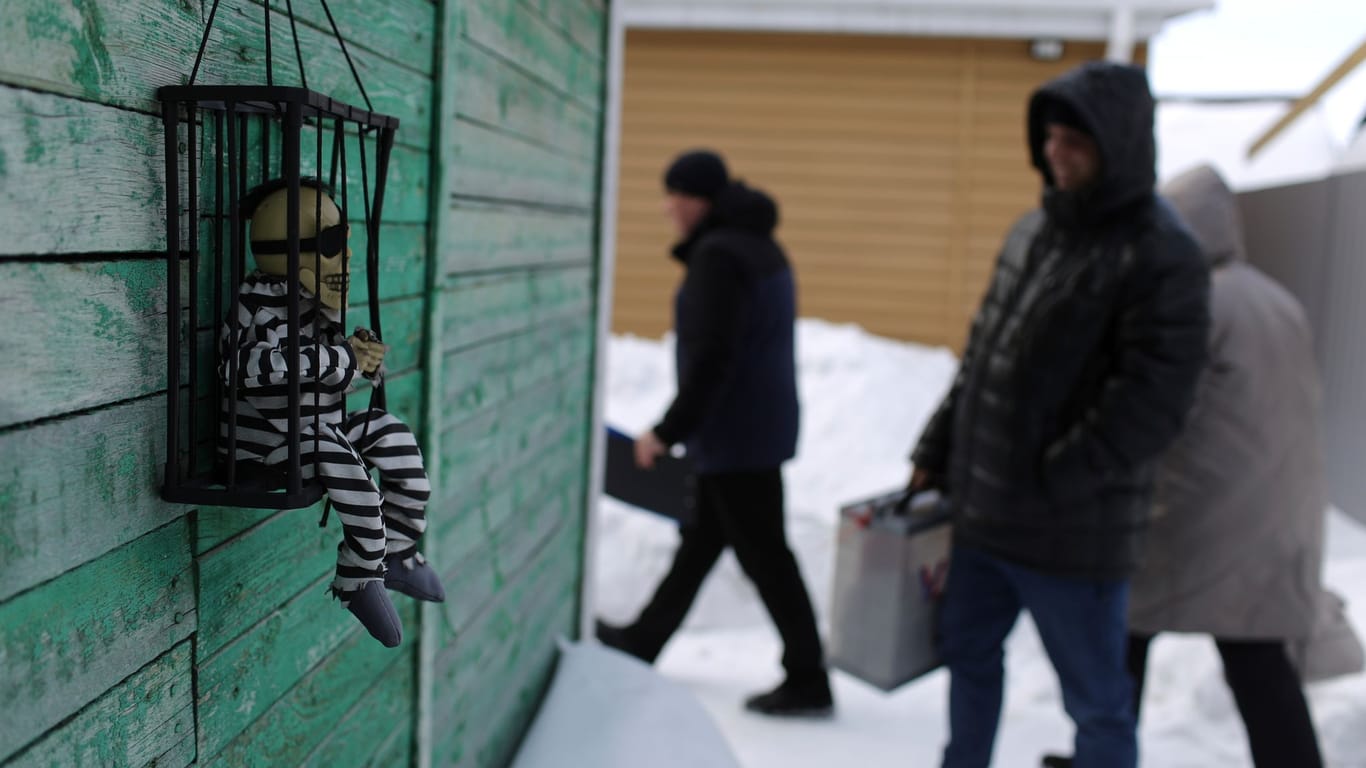 Eine Figur in Häftlingskleidung sitzt in einem Käfig, der an einem Gebäude angebracht ist, an dem russische Wahlhelfer vorbeilaufen.