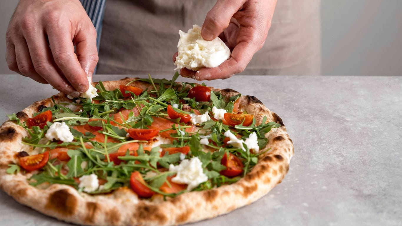 Mann belegt eine Pizza (Symbolfoto): Eine neue Pizzeria lockt mit edlen Zutaten.