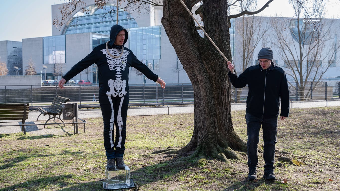 07.03.2024, Berlin, Deutschland, Europa - Der deutsche Klimaaktivist Wolfgang Wolli Metzeler-Kick traegt ein Skelettkostuem und steht mit einer Schlinge um den Hals auf einem schmelzenden Eisblock neben einem Baum, waehrend im Hintergrund das Bundeskanzleramt zu sehen ist.