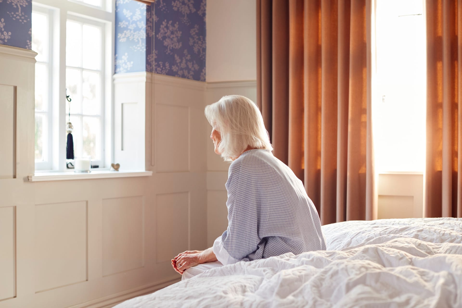 Rückansicht einer älteren Frau, die auf ihrem Bett sitzt.