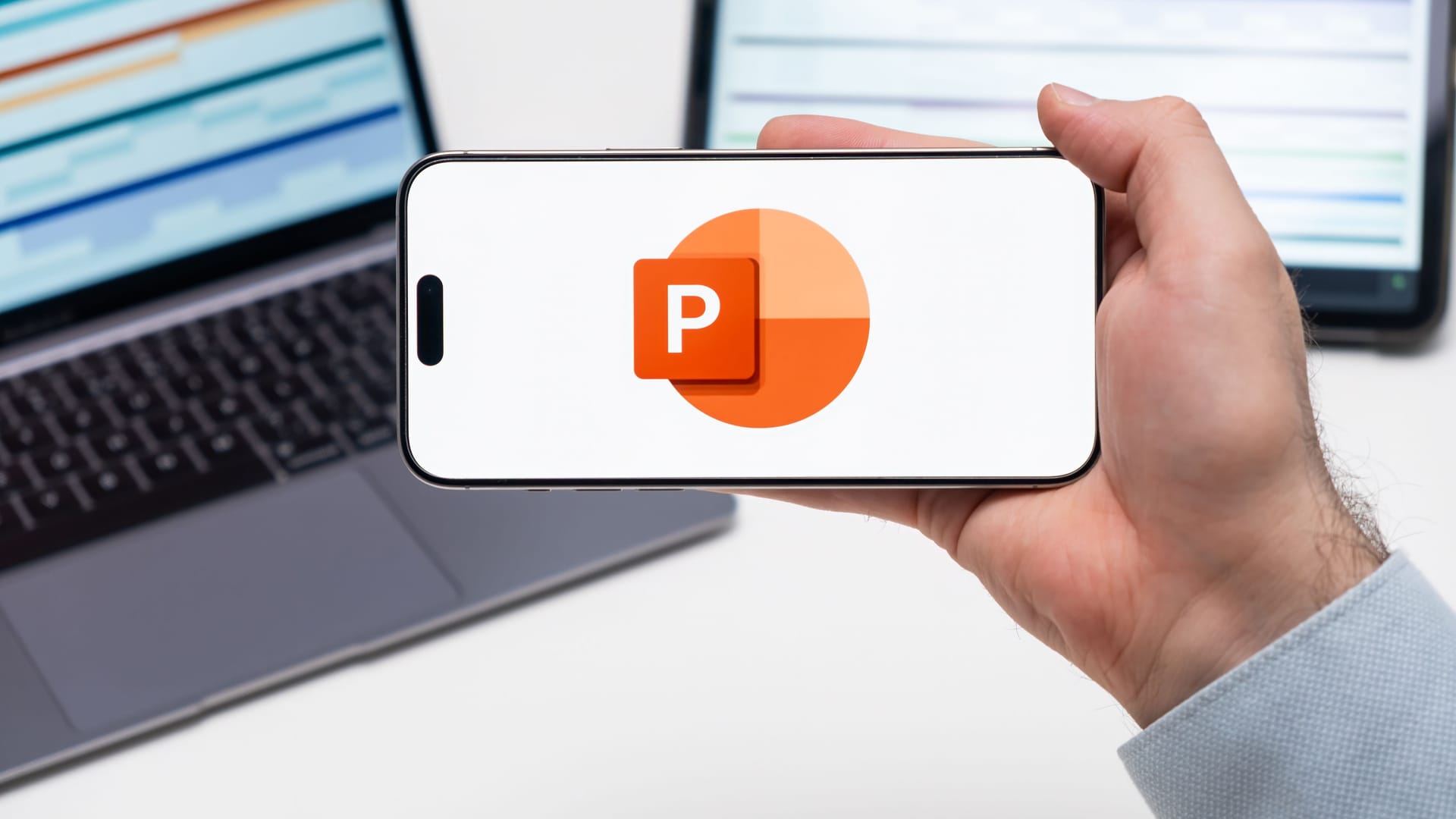 Powerpoint logo on the screen of a smartphone in mans hand with laptop and tablet with graphical diagrams Powerpoint logo on the screen of a smartphone in mans hand with laptop and tablet with graphical diagrams