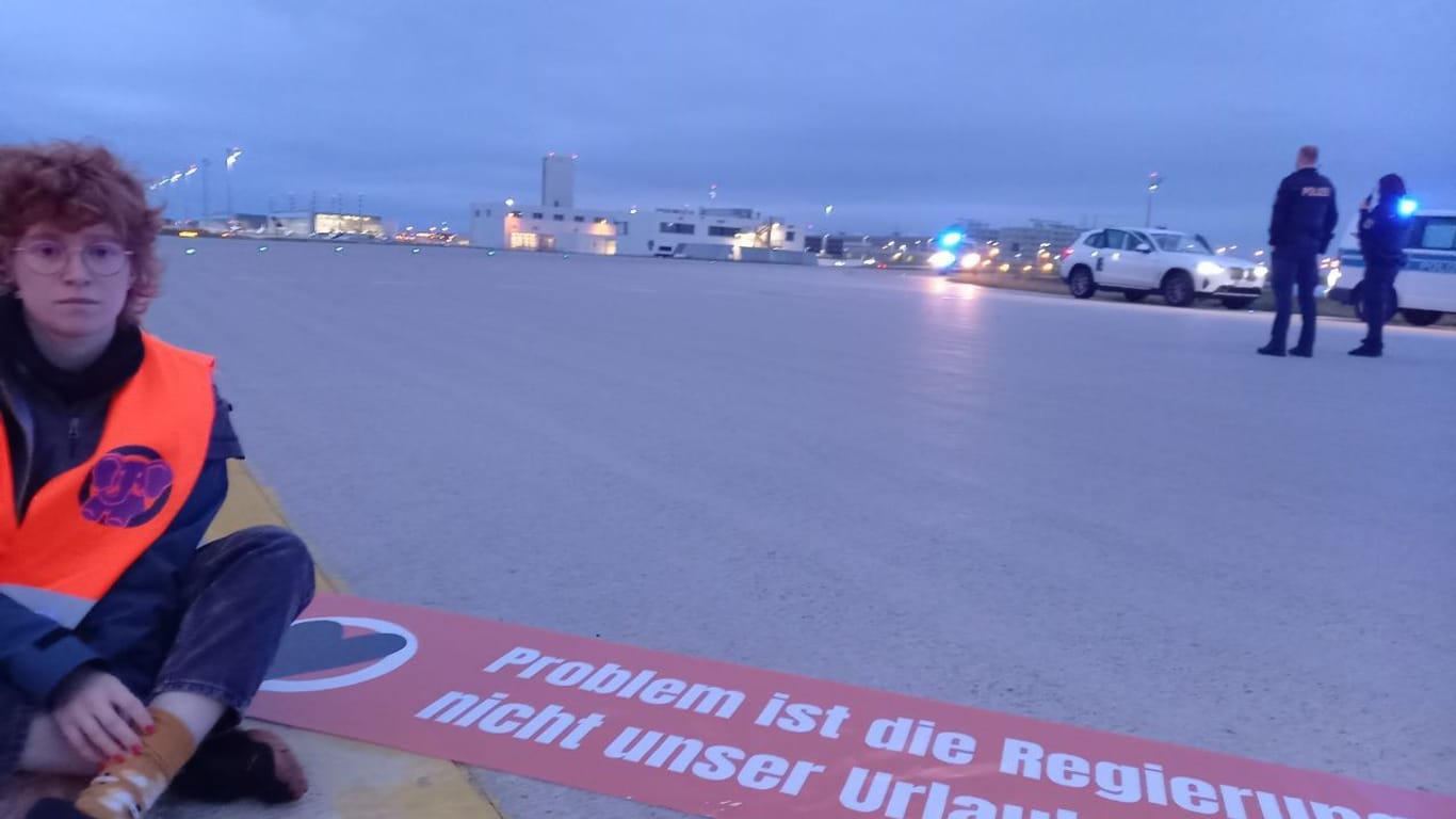 Ein Mitglied der Letzten Generation auf dem Asphalt in München mit einem Banner: "Problem ist die Regierung, nicht unser Urlaub."