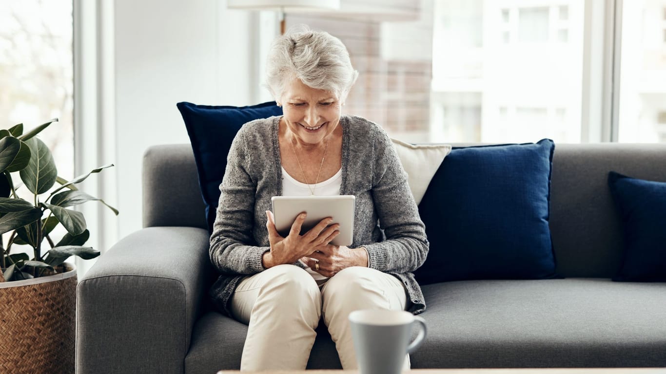 Ältere Frau blickt freudig auf ein Tablet: Wer weiß wie, kann sich mit ETFs entspannt eine Zusatzrente aufbauen.