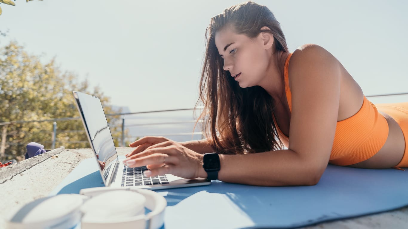 Eine Frau liegt am Strand und arbeitet am Laptop (Symbolbild): Das Konzept "Workation" verbindet den Job mit Urlaub.