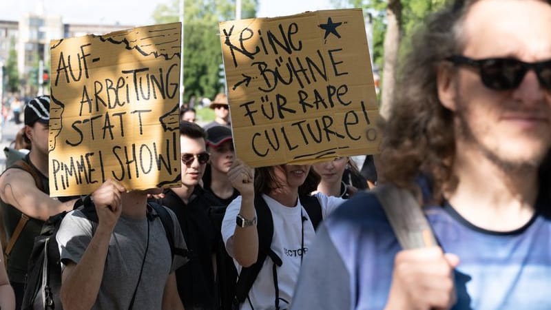 Demonstranten stehen anlässlich einer Kundgebung auf dem Jorge-Gomondai-Platz und halten Schilder mit der Aufschrift "Aufarbeitung statt Pimmelshow" und "Keine Bühne für Rape Culture".