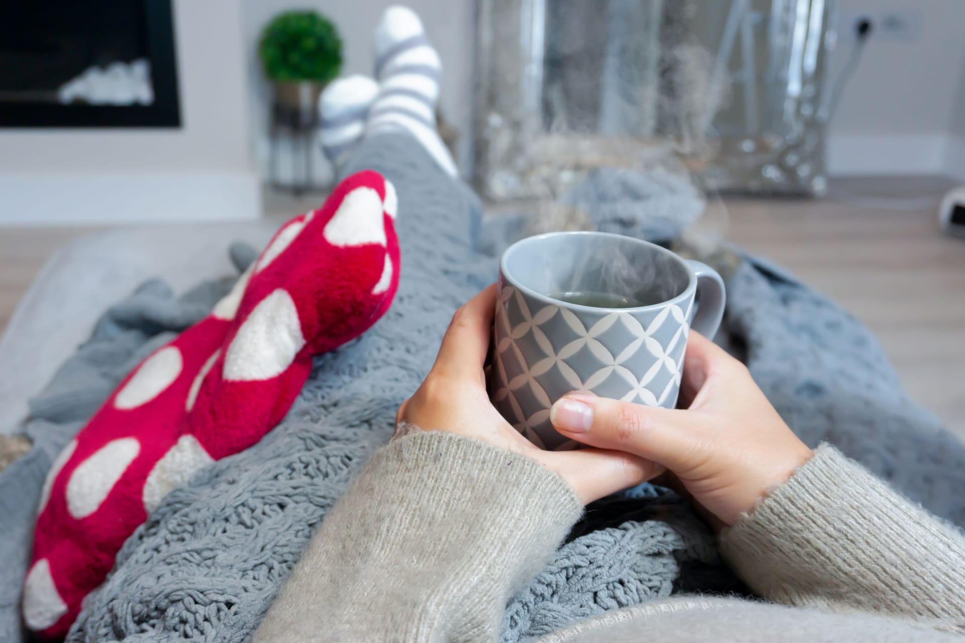 Eine Person liegt mit einer Wärmflasche auf dem Sofa und hält eine Tasse Tee in der Hand.