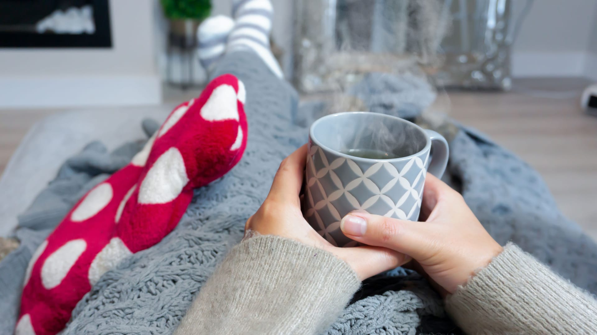 Eine Person liegt mit einer Wärmflasche auf dem Sofa und hält eine Tasse Tee in der Hand.