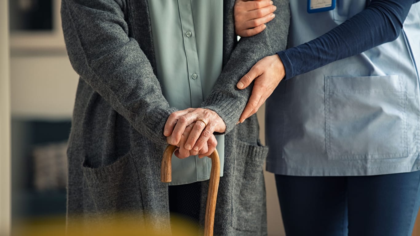 Eine ältere Person mit einem Gehstock erhält Unterstützung durch eine Pflegekraft.