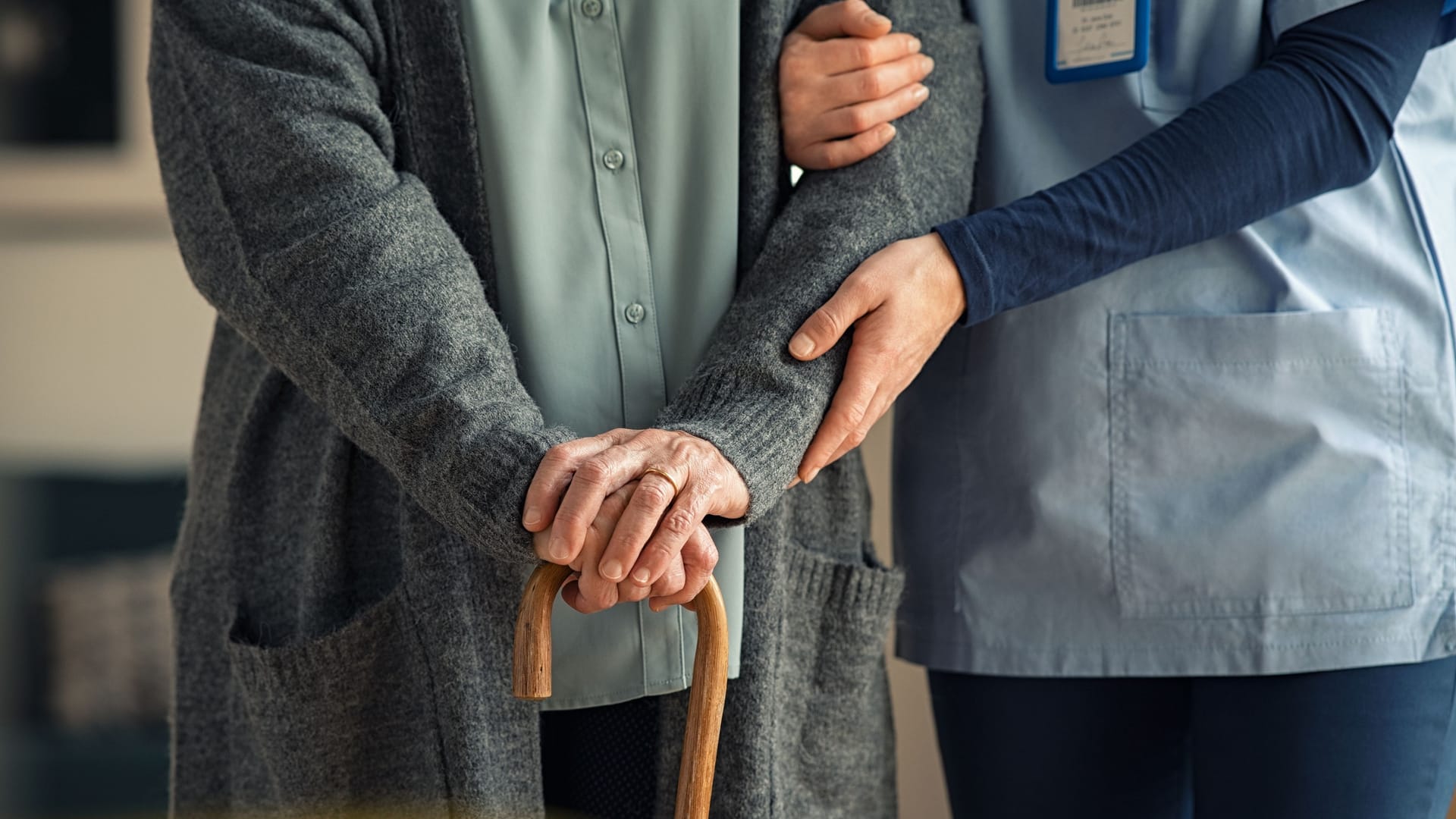 Eine ältere Person mit einem Gehstock erhält Unterstützung durch eine Pflegekraft.
