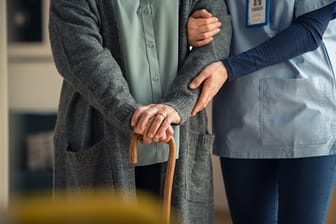 Eine ältere Person mit einem Gehstock erhält Unterstützung durch eine Pflegekraft.