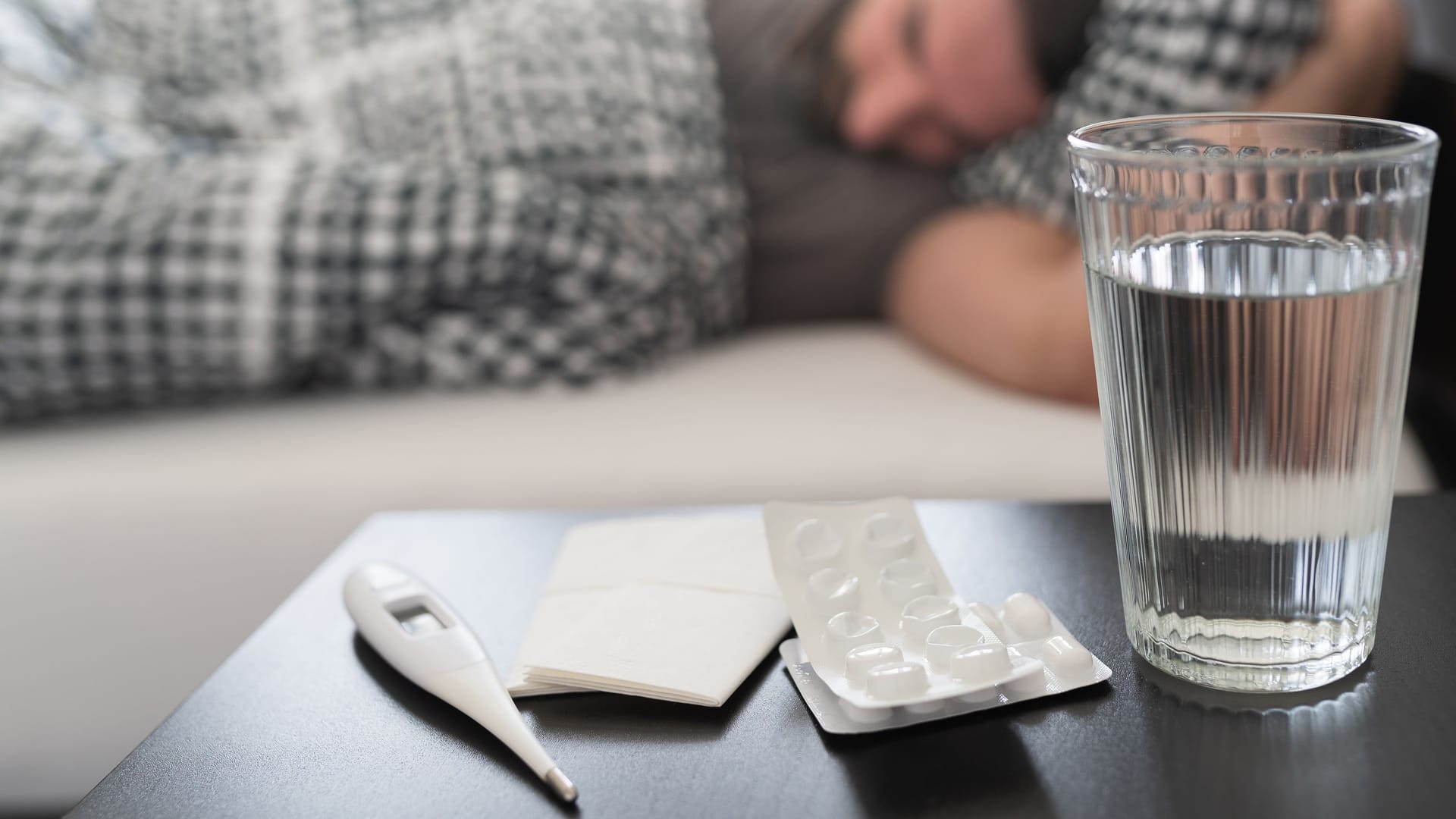 Nachttisch mit Wasserglas, Tabletten und Fieberthermometer, im Hintergrund ein im Bett liegender Mann.