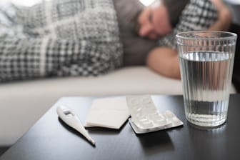 Nachttisch mit Wasserglas, Tabletten und Fieberthermometer, im Hintergrund ein im Bett liegender Mann.