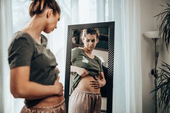 Eine Frau schaut in den Spiegel und befühlt ihren Bauch.