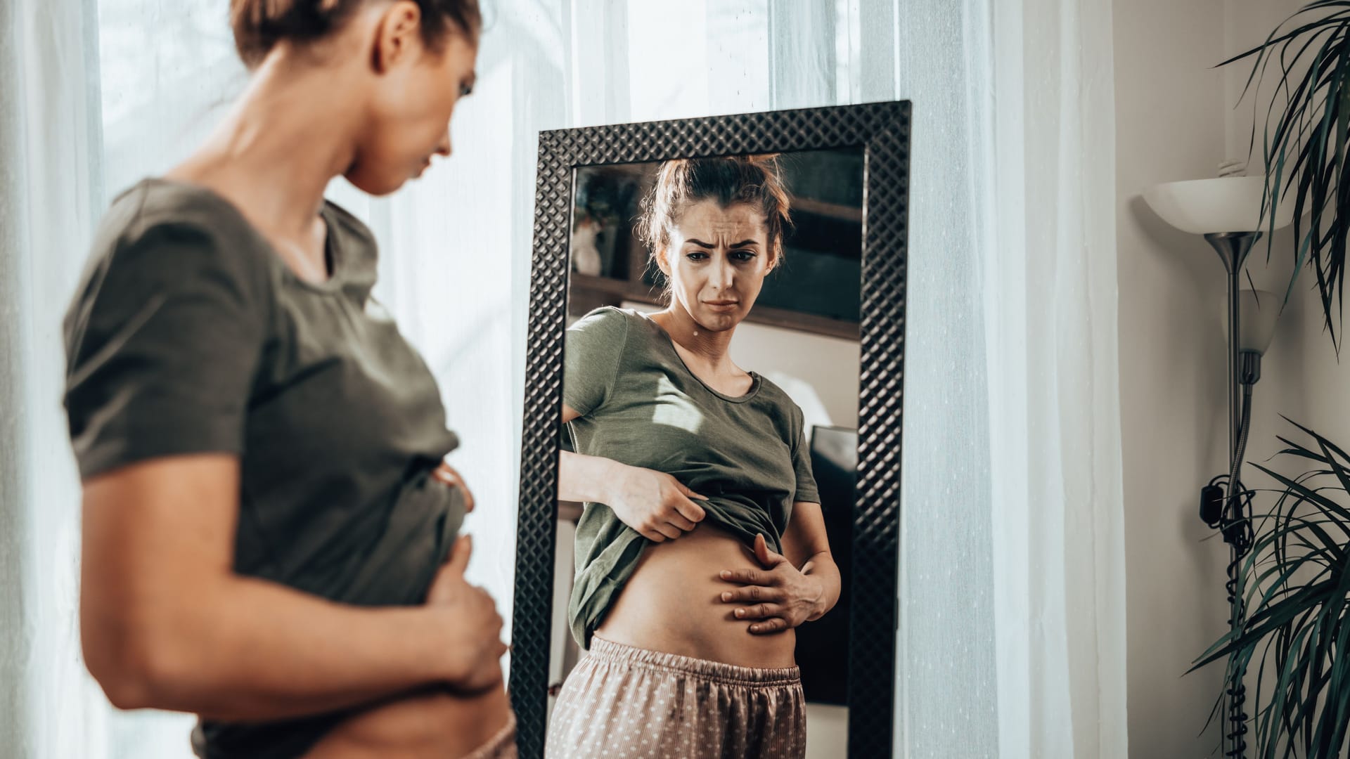 Eine Frau schaut in den Spiegel und befühlt ihren Bauch.