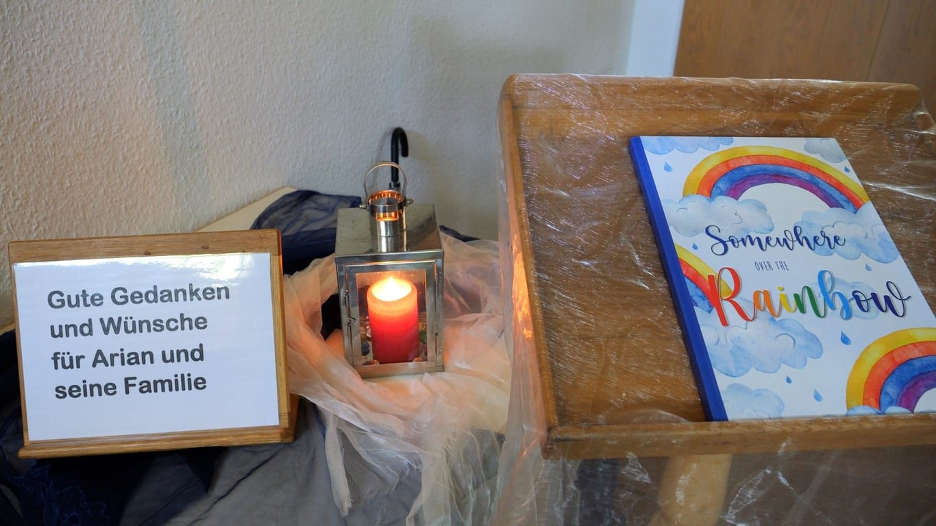 Ein Schild steht neben einer Kerze und ein Buch mit einem Regenbogen auf dem Umschlag liegt daneben: Die Kirche in Elm will Platz zum Trauern geben.
