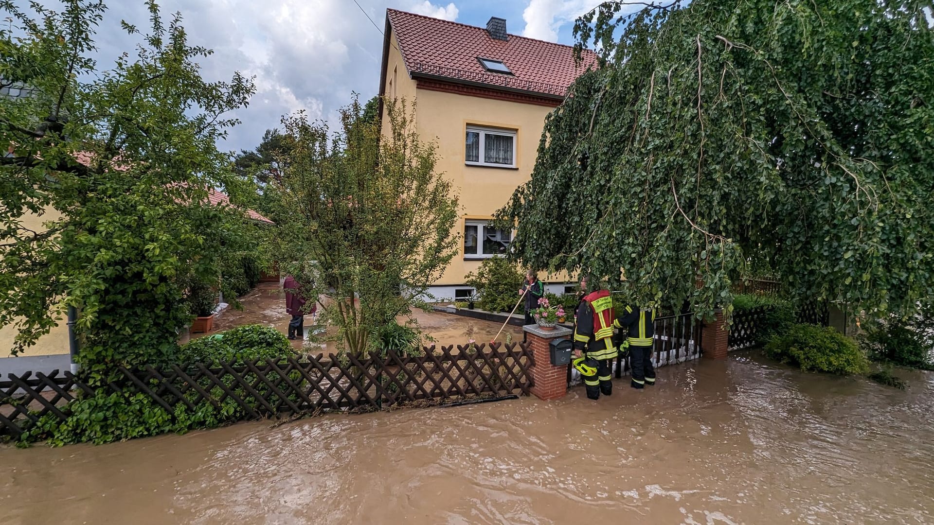 Sachsen, Markkleeberg: Mitarbeiter der Feuerwehr unterhalten sich mit Anwohnern eines im Wasser stehenden Hauses.