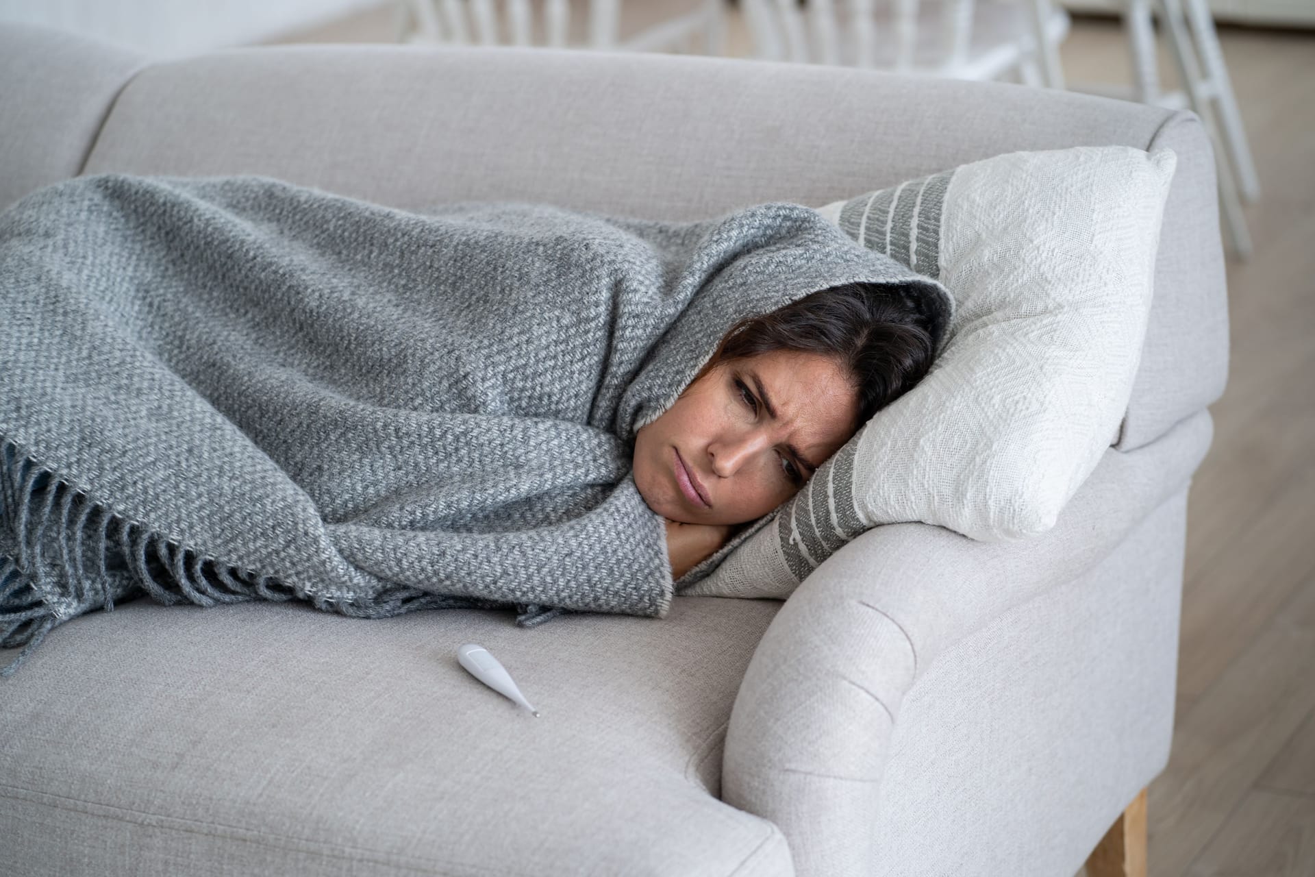 Frau liegt mit Wolldecke auf dem Sofa, neben ihr ein Fieberthermometer