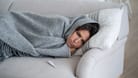 Frau liegt mit Wolldecke auf dem Sofa, neben ihr ein Fieberthermometer