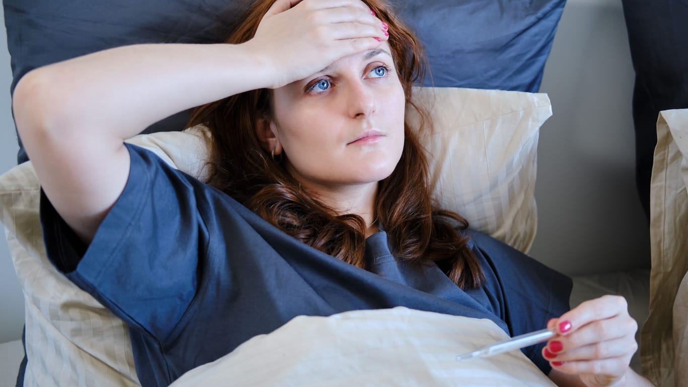 Frau liegt im Bett und fasst sich mit einer Hand an die Stirn, in der anderen Hand hält sie ein Fieberthermometer