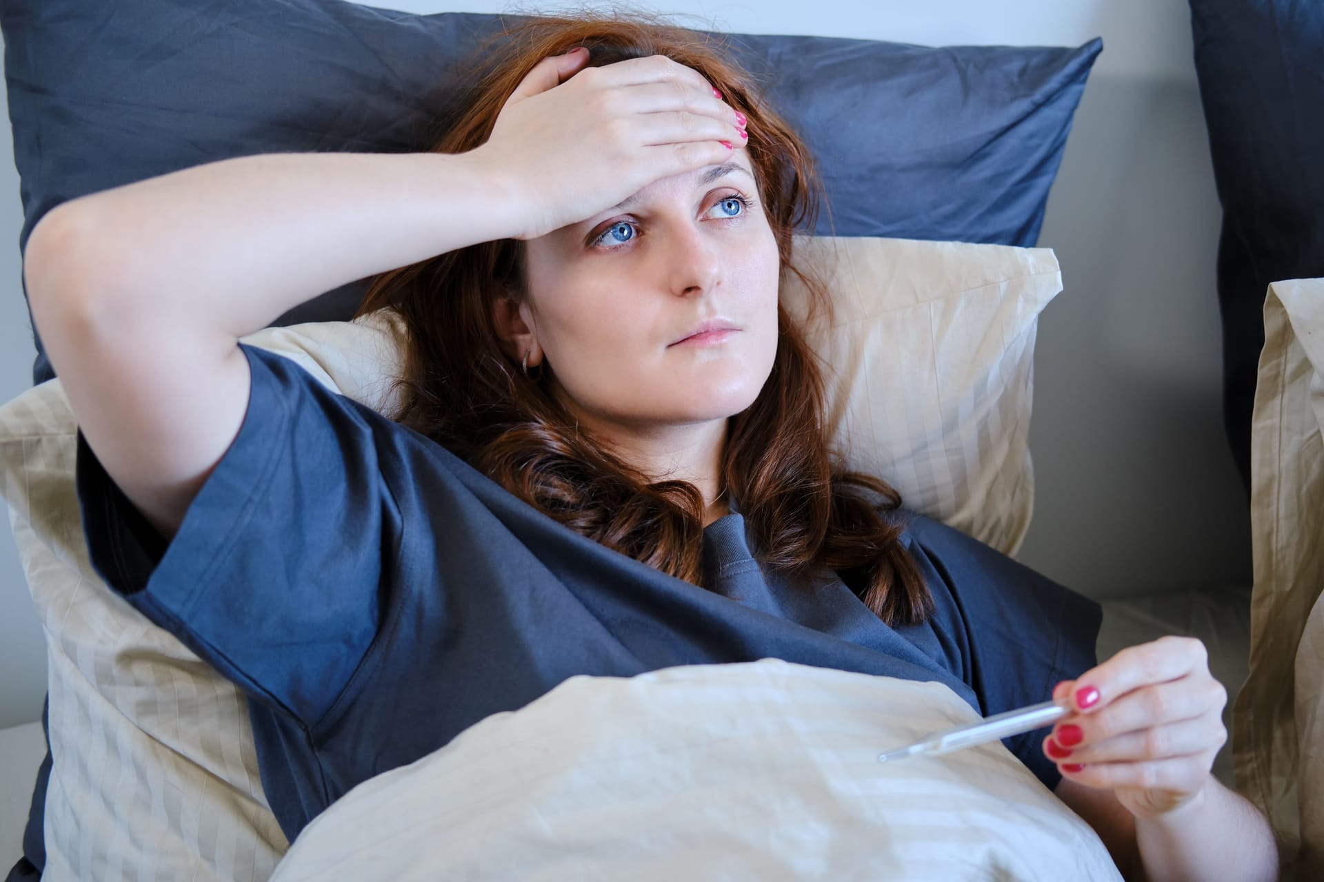 Frau liegt im Bett und fasst sich mit einer Hand an die Stirn, in der anderen Hand hält sie ein Fieberthermometer