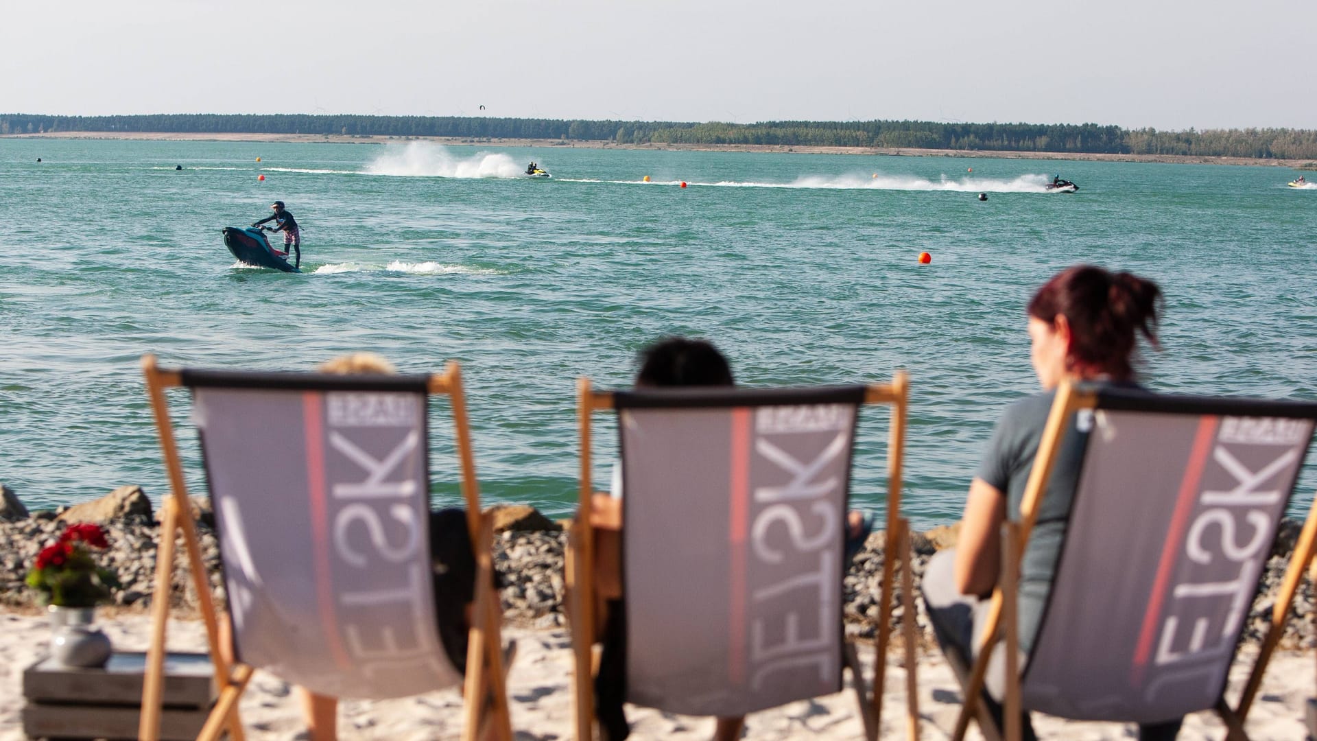 Drei Frauen sitzen auf Klappstühlen und beobachten Jetskis auf dem Partwitzer See.